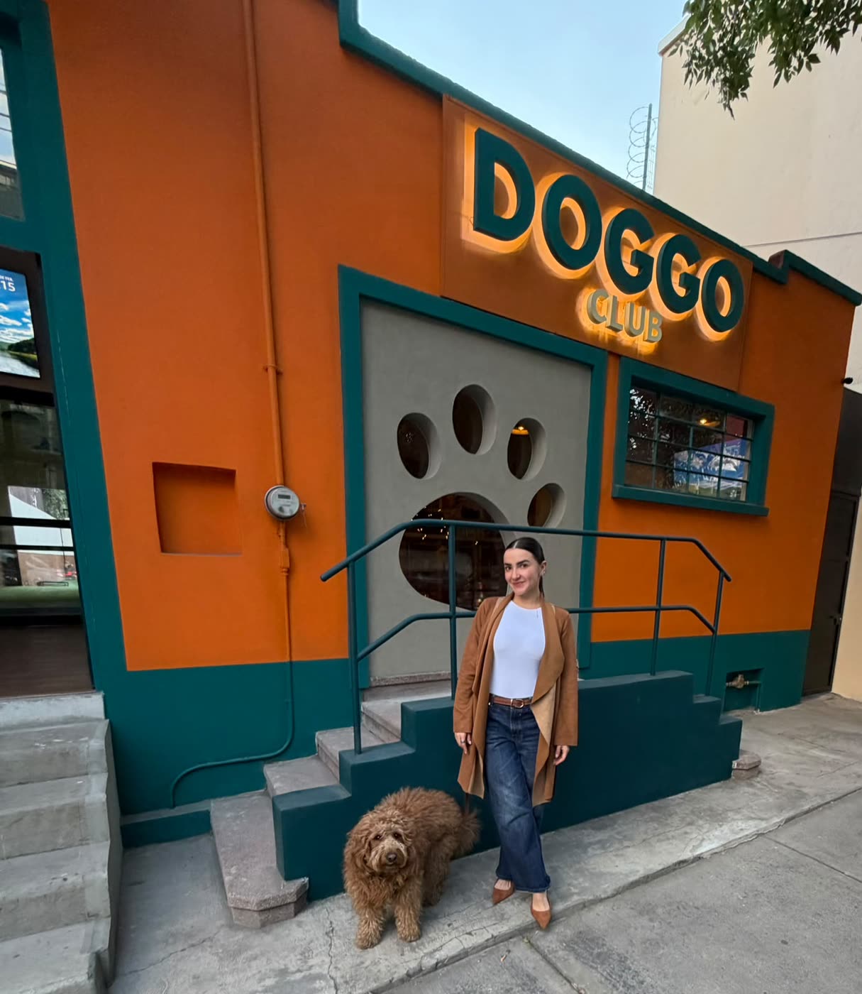 Fachada de Doggo Club al atardecer: letrero luminoso sobre muro naranja y verde, con la huella troquelada y una mujer acompañada de un perro café en la entrada.